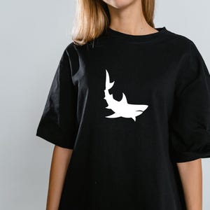 Puede incluir: Una camiseta negra con un gr&aacute;fico de silueta de tibur&oacute;n blanco.