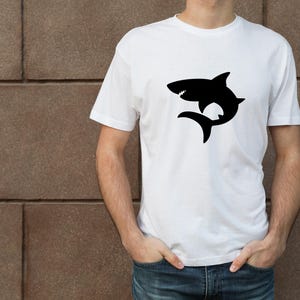 Puede incluir: Una camiseta blanca con una silueta negra de un tibur&oacute;n en la parte delantera.