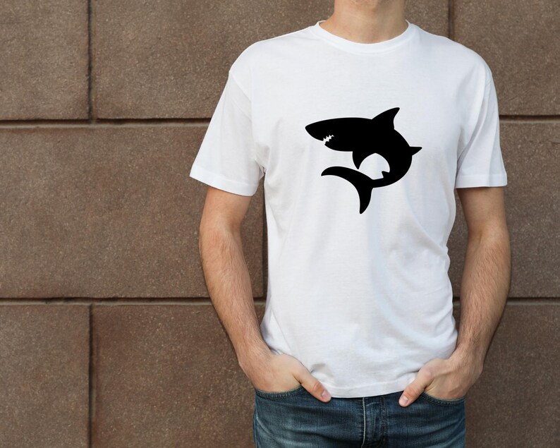Puede incluir: Una camiseta blanca con una silueta negra de un tibur&oacute;n en la parte delantera.
