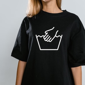 Może przedstawiać: Czarny T-shirt z białym symbolem prania ręcznego. Symbol przedstawia rękę w misce z wodą.