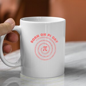 K&ouml;nnte beinhalten: Wei&szlig;e Keramiktasse mit einem rosafarbenen Design, auf dem "Geboren am Pi-Tag" steht und eine Spirale aus den Ziffern von Pi mit einem gro&szlig;en Pi-Symbol in der Mitte zu sehen ist.