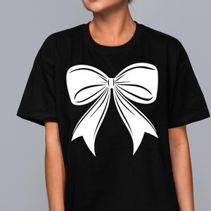 May include: Black t-shirt with a large white bow graphic printed on the front. The bow is a simple, elegant design. The t-shirt is a casual item of clothing.