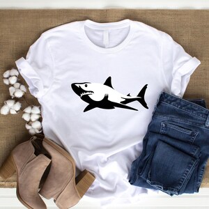 Puede incluir: Camiseta blanca con una silueta negra de un tibur&oacute;n en la parte delantera. La camiseta est&aacute; sobre una superficie marr&oacute;n con un par de botines marrones y unos vaqueros azules.