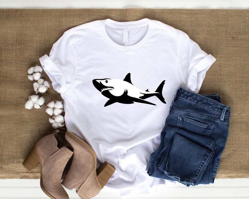 Puede incluir: Camiseta blanca con una silueta negra de un tibur&oacute;n en la parte delantera. La camiseta est&aacute; sobre una superficie marr&oacute;n con un par de botines marrones y unos vaqueros azules.
