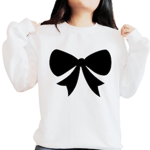 May include: White sweatshirt featuring a large black bow graphic. The long-sleeved top is made of a soft material, suitable for casual wear. The bow is a simple, elegant design, perfect for everyday wear.