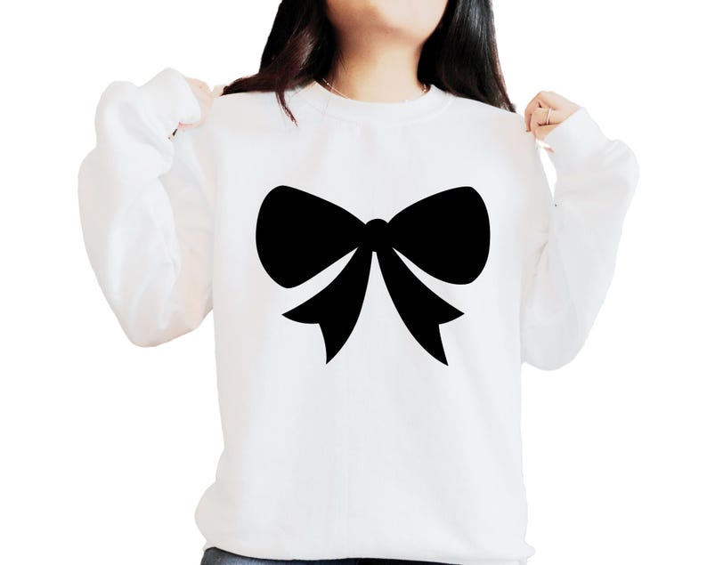 May include: White sweatshirt featuring a large black bow graphic. The long-sleeved top is made of a soft material, suitable for casual wear. The bow is a simple, elegant design, perfect for everyday wear.