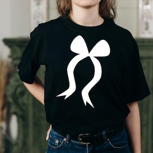 May include: Black t-shirt with a large white bow graphic. The bow design has a classic shape with flowing ribbons. The shirt is made of a soft material and has short sleeves.