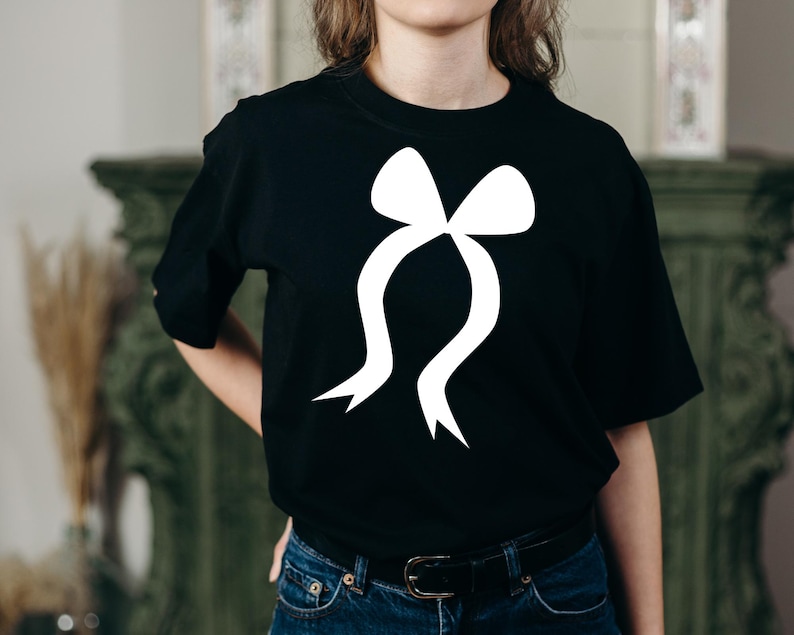 May include: Black t-shirt with a large white bow graphic. The bow design has a classic shape with flowing ribbons. The shirt is made of a soft material and has short sleeves.