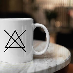 Może przedstawiać: Biały ceramiczny kubek z czarnym symbolem tr&oacute;jkąta z przekreśloną przekątną. Kubek stoi na białym marmurowym blacie.