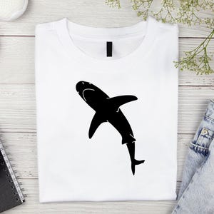 Puede incluir: Una camiseta blanca con una silueta negra de un tibur&oacute;n en la parte delantera.