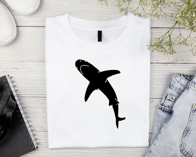 Puede incluir: Una camiseta blanca con una silueta negra de un tibur&oacute;n en la parte delantera.