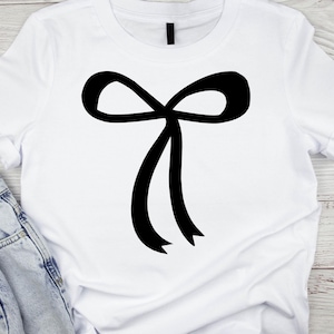 May include: White t-shirt with a large black bow graphic. The bow is centrally placed and has two long, flowing ribbons. The shirt is made of a soft material, suitable for casual wear.