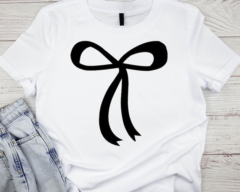 May include: White t-shirt with a large black bow graphic. The bow is centrally placed and has two long, flowing ribbons. The shirt is made of a soft material, suitable for casual wear.