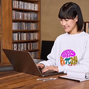 K&ouml;nnte beinhalten: Eine Frau tr&auml;gt ein wei&szlig;es Sweatshirt mit einer bunten Grafik, auf der "Happy Pi 3.14 Day" steht, und arbeitet an einem Laptop an einem Holztisch.