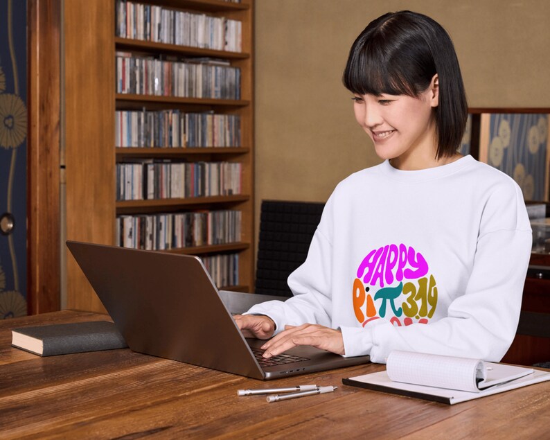 K&ouml;nnte beinhalten: Eine Frau tr&auml;gt ein wei&szlig;es Sweatshirt mit einer bunten Grafik, auf der "Happy Pi 3.14 Day" steht, und arbeitet an einem Laptop an einem Holztisch.