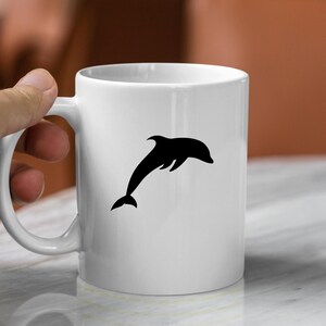 Puede incluir: Taza de cer&aacute;mica blanca con una silueta negra de un delf&iacute;n saltando fuera del agua.