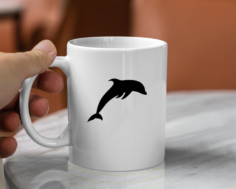 Puede incluir: Taza de cer&aacute;mica blanca con una silueta negra de un delf&iacute;n saltando fuera del agua.