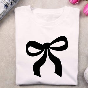May include: A white t-shirt with a large, black bow graphic. The bow is centrally placed and has a traditional design. The t-shirt is made from a soft fabric, suitable for everyday wear.