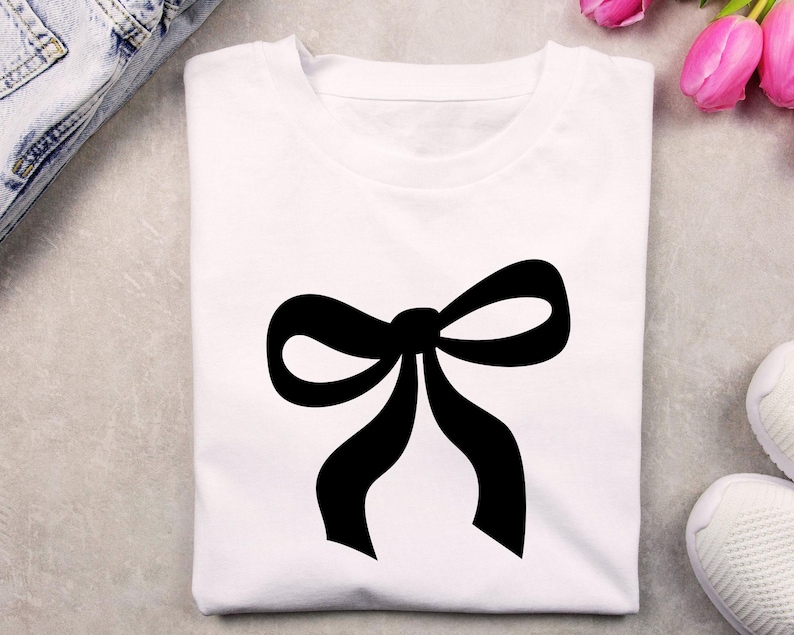 May include: A white t-shirt with a large, black bow graphic. The bow is centrally placed and has a traditional design. The t-shirt is made from a soft fabric, suitable for everyday wear.