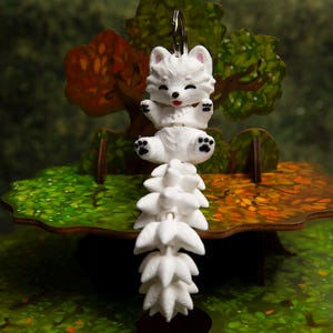 May include: A white fox keychain featuring a small, seated fox figure with a long, articulated, multi-segmented tail. The fox has black eyes, a black nose, a small red tongue, and black paw pads on its feet. A silver metal ring is attached to the top.