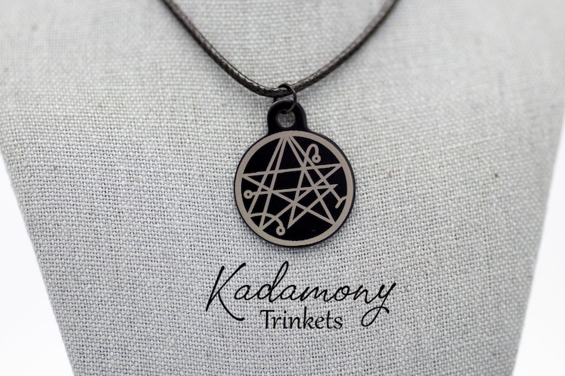 May include: A black pendant necklace with a silver five-pointed star with a circle around it. The pendant is on a black cord. The text "Kadomony Trinkets" is visible below the pendant.