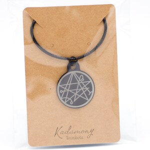 May include: A black necklace with a silver pendant featuring a five-pointed star with intricate lines. The pendant is on a brown card with the text "Kadamony Trinkets".