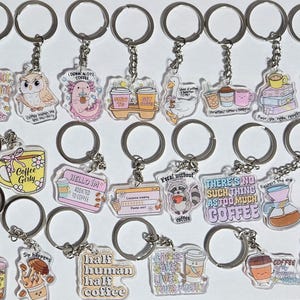 May include: A collection of twenty acrylic keychains featuring various coffee-themed illustrations and text, including phrases like 'Probably on my way to get coffee', 'Coffee Girly', 'Feral without coffee', and 'There's no such thing as too much coffee'.