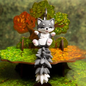 May include: A gray and white articulated fox keychain featuring a small, smiling fox figure with pink paw pads and a long, segmented tail that hangs down, attached to a silver metal keyring loop for secure attachment to bags or keys.