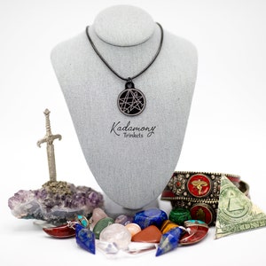 May include: A black necklace with a circular pendant featuring a geometric design. The necklace is displayed on a gray mannequin. The image also includes a small silver sword, a cluster of amethyst crystals, and a variety of colorful gemstones. The text "Kadomony Trinkets" is visible below the necklace.
