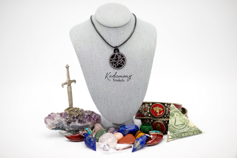 May include: A black necklace with a circular pendant featuring a geometric design. The necklace is displayed on a gray mannequin. The image also includes a small silver sword, a cluster of amethyst crystals, and a variety of colorful gemstones. The text "Kadomony Trinkets" is visible below the necklace.