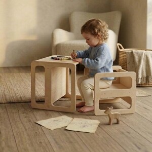 May include: A light-colored wooden children's table and chair set. The table has a square top and open sides. A child is seated at the chair, drawing with crayons. A small wooden deer figurine is on the floor.