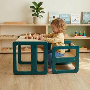 May include: A child sits at a teal wooden table and chair set. The table has a light wood top. The child is playing with wooden toys. Shelves with books and toys are in the background. A potted plant is also visible.
