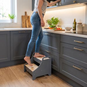 May include: A gray wooden step stool with two steps and natural wood treads. The step stool is in a kitchen setting, with a person using it to reach a cabinet. The kitchen has gray cabinets and a light countertop.