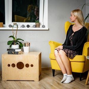 May include: A light-colored wooden side table with circular cutouts of varying sizes. The table is next to a yellow armchair. A woman is sitting in the armchair, wearing a black cardigan and a floral dress. A plant and books are on the table.