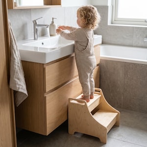 May include: A light-colored wooden two-step stool with side handles and a smooth finish. The stool features a wide base and two horizontal steps, designed to provide elevation for reaching bathroom sinks or countertops in a home environment.