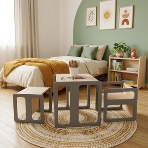 May include: A children's furniture set consisting of a square table and two matching chairs, all featuring a light wood finish with grey side panels. A white cup filled with colored pencils sits on the table surface, which is set on a round rug.