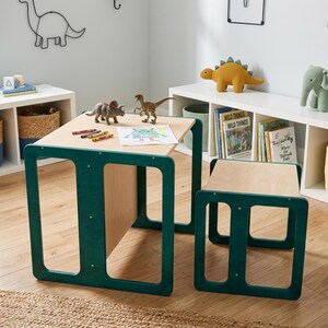 May include: A light wood children's table and matching bench set with dark green painted frames. The table surface holds a drawing of a monster, three crayons, and two plastic dinosaur figurines. The furniture features a simple, open-frame design.