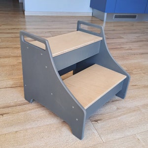 May include: A two-step grey wooden stool with a light wood top. The stool has rounded edges and a simple design.