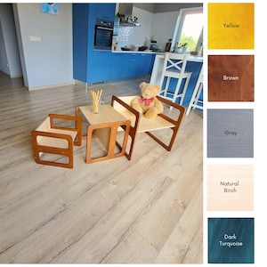 May include: A child's wooden table and chair set with a natural birch tabletop and brown frame. The set includes a small table and two chairs. A teddy bear sits on one of the chairs. The set is on a light wood floor.