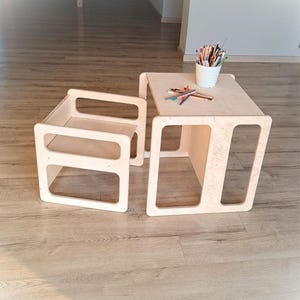 May include: A light-colored wooden children's table and chair set. The table has a square top with a lower shelf, and the chair has a back and seat. A white cup with colored pencils sits on the table. The set is designed with cut-out rectangular shapes.