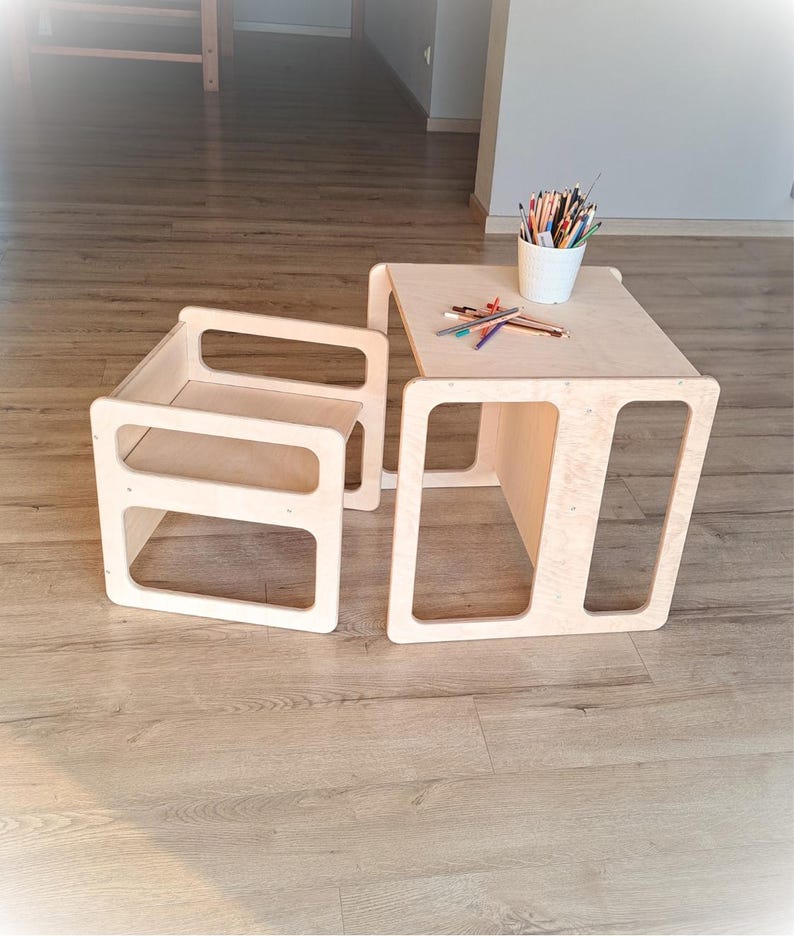 May include: A light-colored wooden children's table and chair set. The table has a square top with a lower shelf, and the chair has a back and seat. A white cup with colored pencils sits on the table. The set is designed with cut-out rectangular shapes.