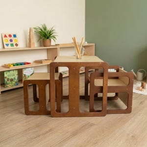 May include: A wooden children's table and two matching chairs with a natural light wood top and dark brown frame. The table features a clear glass jar filled with wooden sticks. The furniture set has a simple, open-frame design with rounded edges.