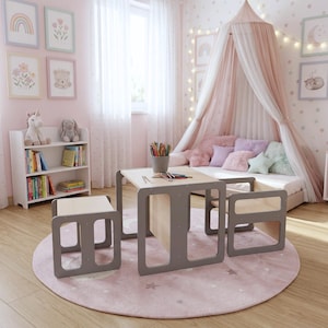 May include: A children's bedroom scene featuring a small table and two chairs with gray frames and light wood tops. A matching bookshelf and a round pink rug with star patterns are also present. The room is decorated with framed art and a canopy bed.