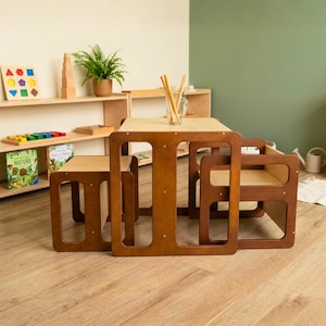 May include: A wooden children's table and two chairs set in a light brown finish. The table has a rectangular top and open sides. The chairs are square with open sides. A shelf with educational toys and books is in the background.