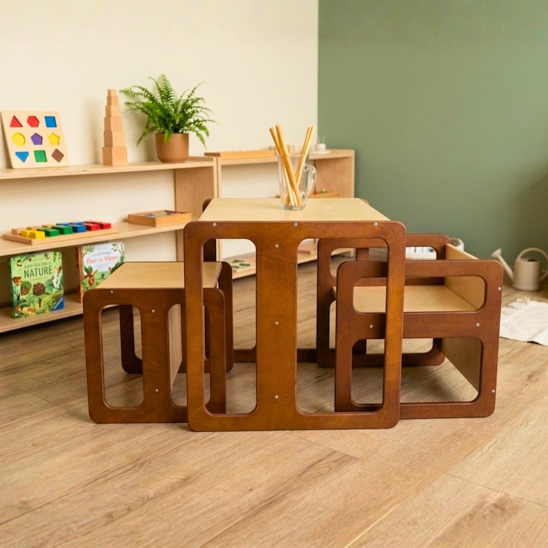 May include: A wooden children's table and two chairs set in a light brown finish. The table has a rectangular top and open sides. The chairs are square with open sides. A shelf with educational toys and books is in the background.