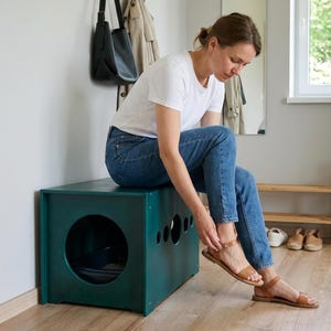 May include: A teal wooden bench with a large circular opening and several smaller holes. The bench is rectangular and appears to be a storage unit. A person is sitting on the bench, wearing blue jeans and brown sandals.