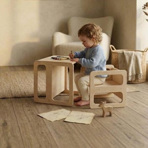 May include: A light-colored wooden children's table and chair set. The table has a square top and open sides. A child is seated at the chair, drawing with crayons. A small wooden deer figurine is on the floor.