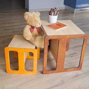 May include: A child's wooden table and chair set. The table and chair have a natural wood top and a brown frame. The chair frame is orange. A teddy bear sits on the chair. A white cup of colored pencils and a notebook are on the table.