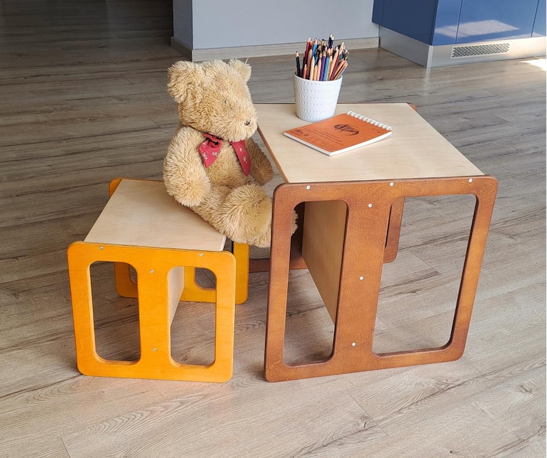 May include: A child's wooden table and chair set. The table and chair have a natural wood top and a brown frame. The chair frame is orange. A teddy bear sits on the chair. A white cup of colored pencils and a notebook are on the table.