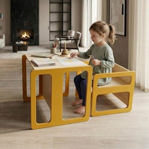 May include: A child's yellow table and chair set. The table has a light wood top and a square frame with cut-out rectangles. The chair has a similar design. The child is drawing on a piece of paper with crayons.